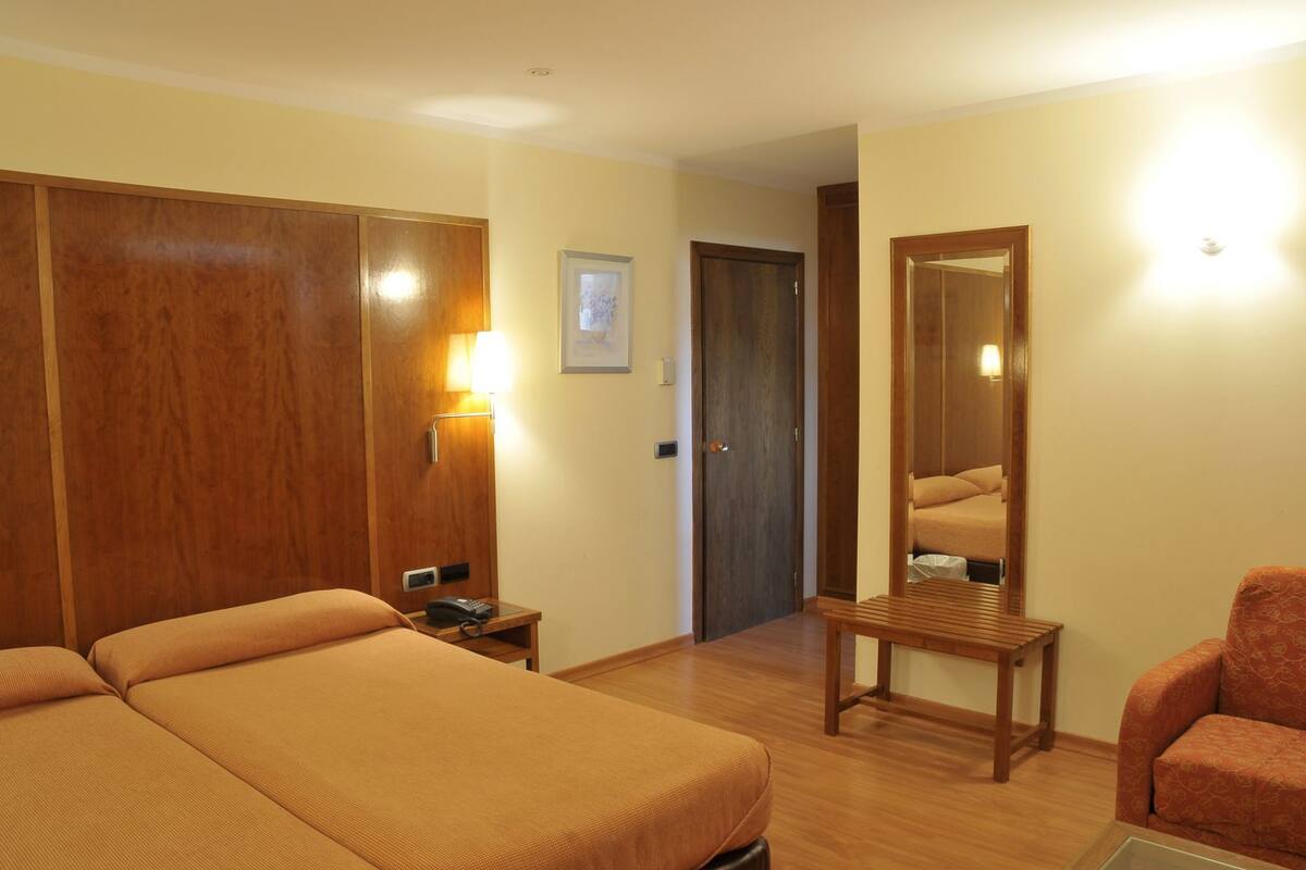 A hotel room with two beds, a wooden wardrobe, a bedside table with a lamp, a mirror, and a small armchair.