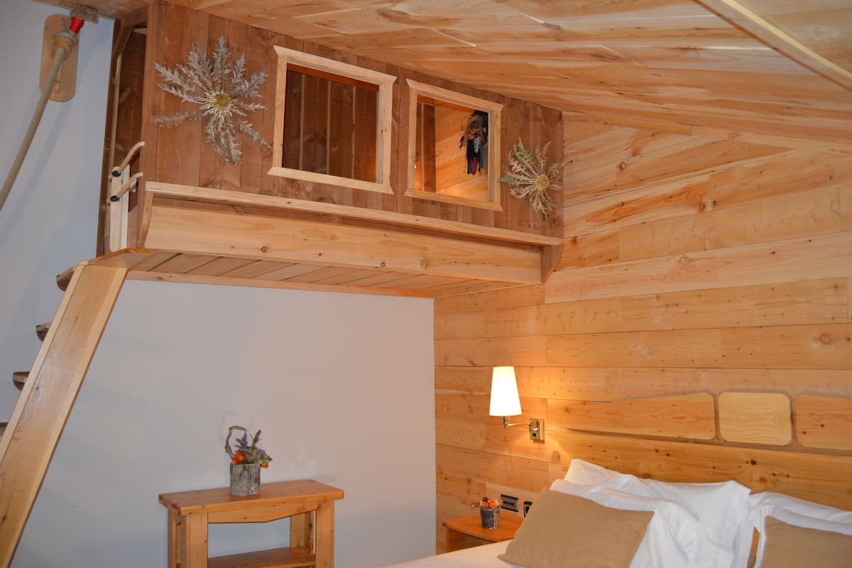 A wooden cabin interior with a bed, a small table with a plant, and a window with a view.