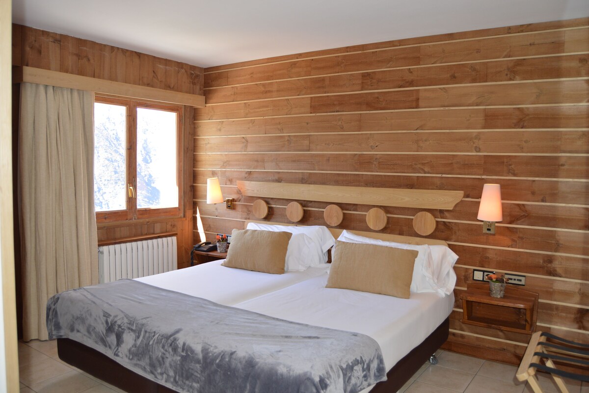 A double bed with a wooden headboard, two bedside lamps, a nightstand, and a window with curtains.