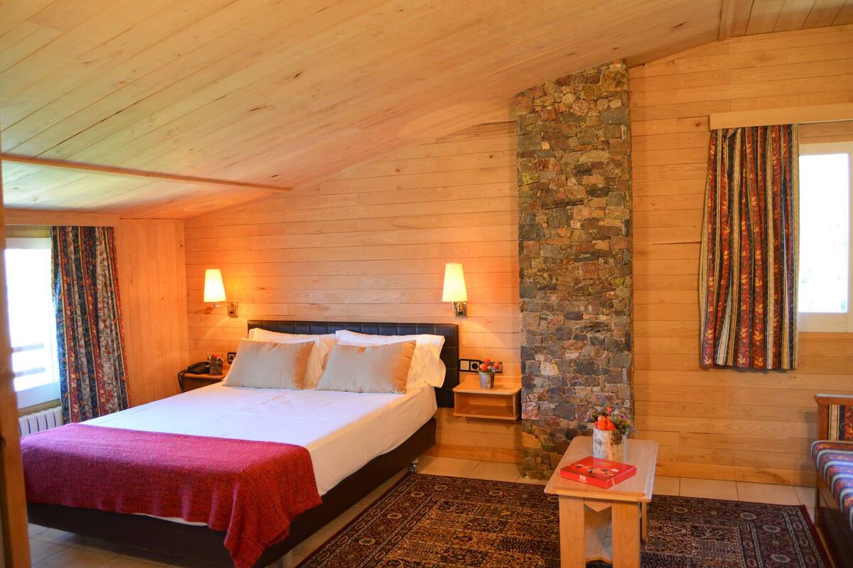 A cozy room with a bed, two bedside lamps, a stone wall, a wooden ceiling, and a window with curtains.