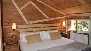 A wooden cabin bedroom with a bed, two bedside lamps, a nightstand, and a window with a view of trees. - Hotel Coma Bella (Sant Julia de Loria)