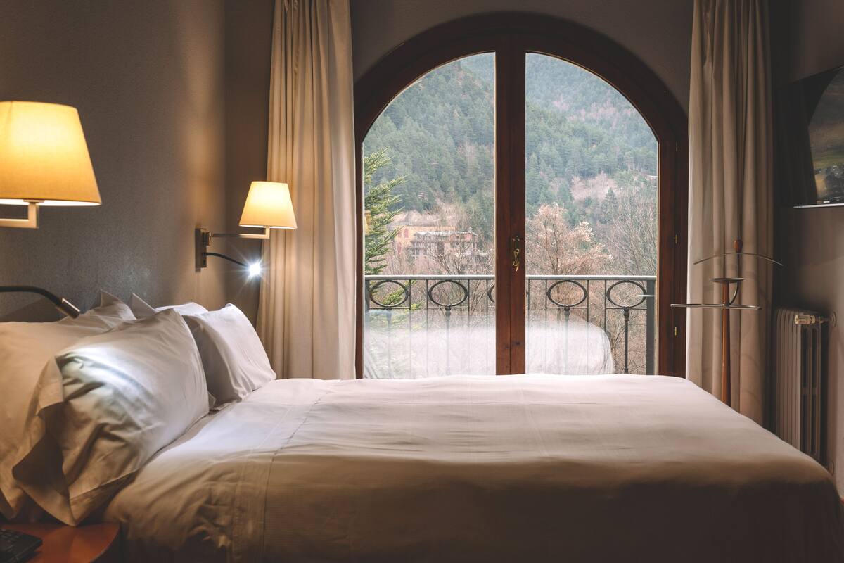 A hotel room with a large bed, two bedside lamps, a view of a balcony with a railing, and a door leading to an outdoor area.