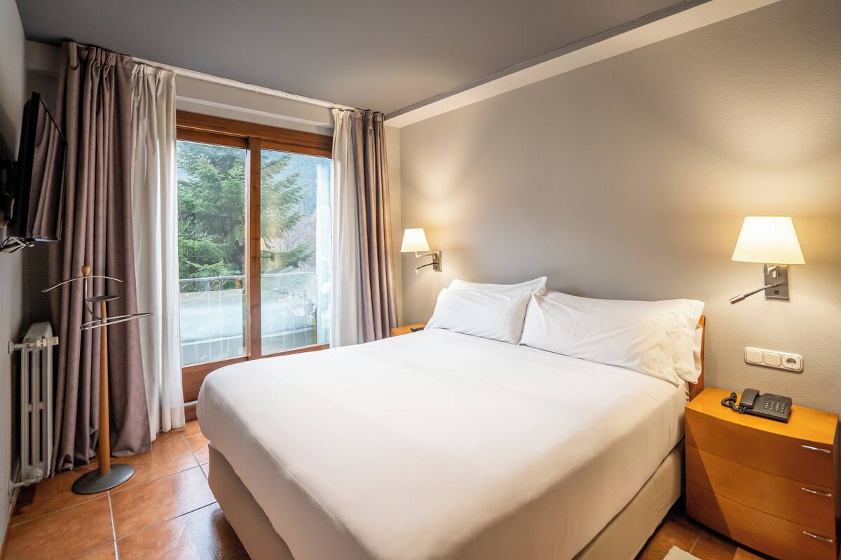 A hotel room with a large bed, two bedside lamps, a wooden nightstand, and a view of greenery outside.