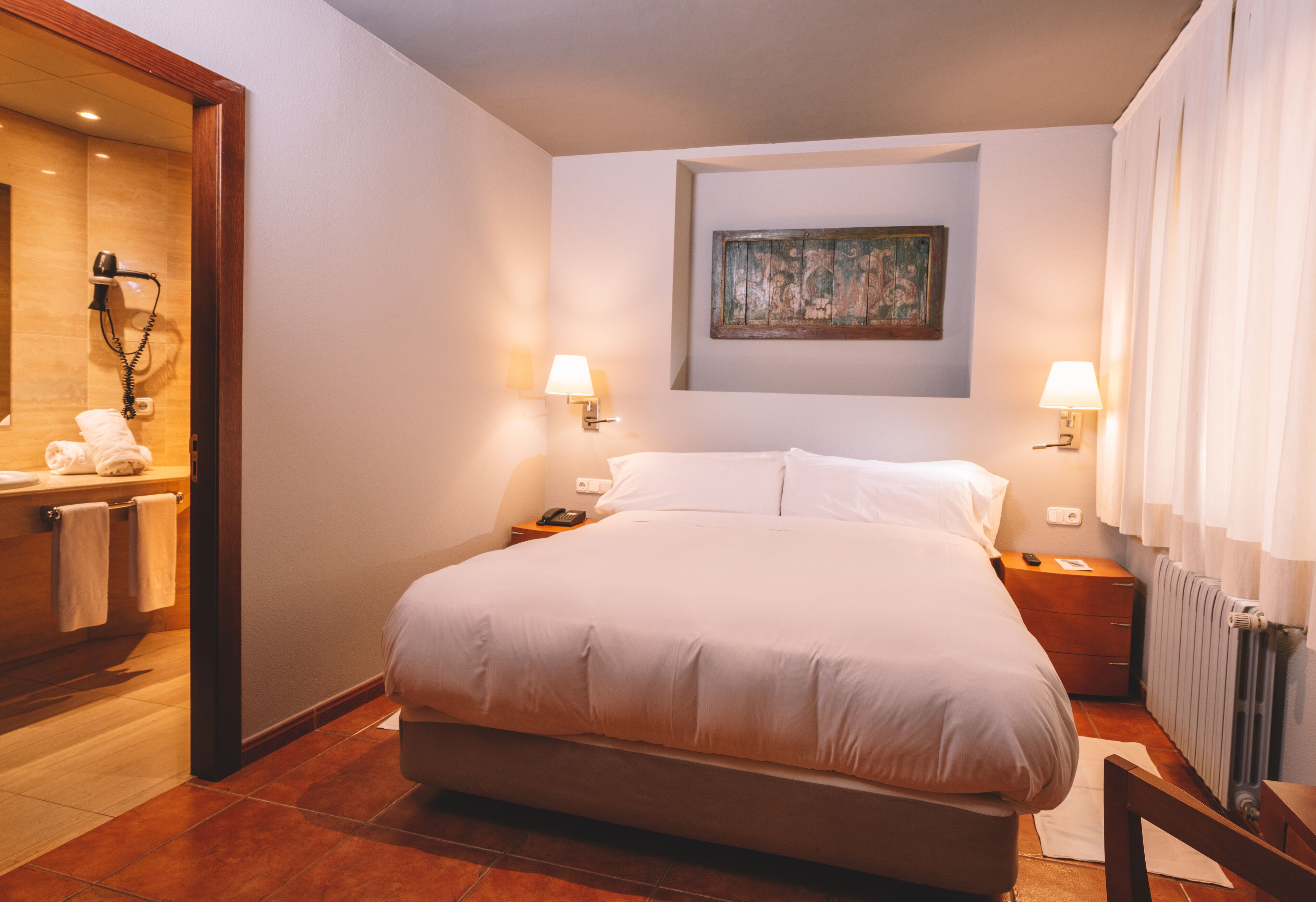 Un dormitorio ordenado con una cama grande, dos mesitas de noche, un tocador, un cuadro colgado en la pared y un baño visible a través de una puerta abierta.
