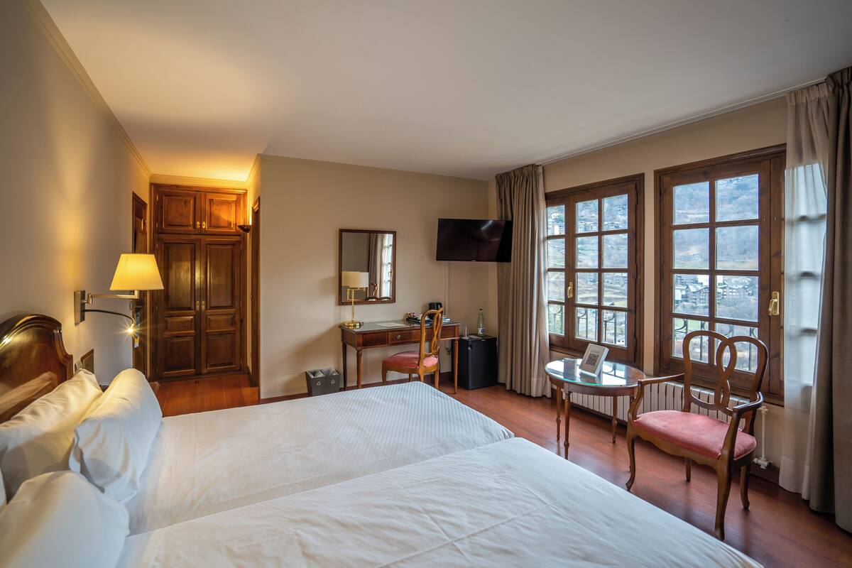 A hotel room with two beds, a desk with a chair, a TV, and large windows.