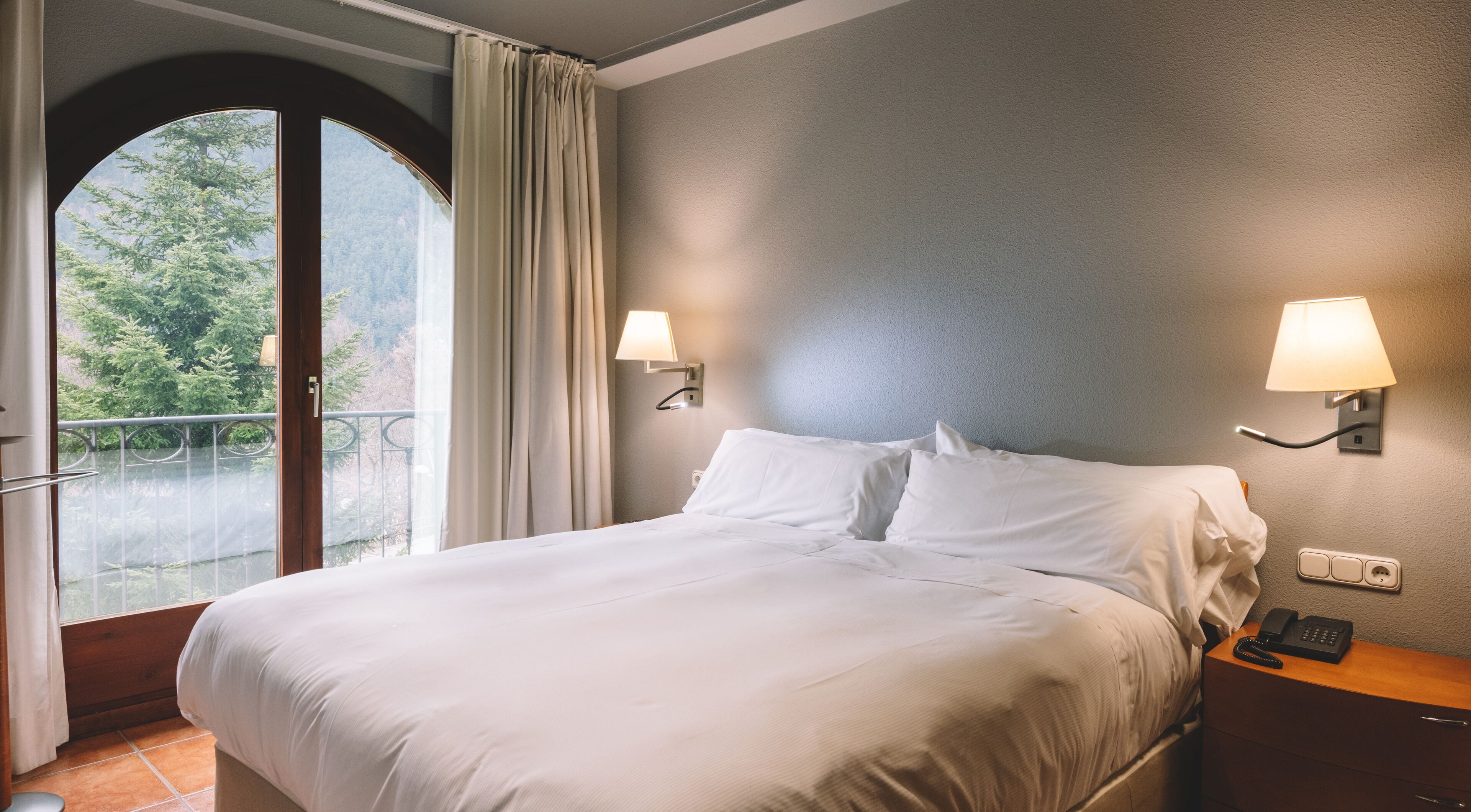 Habitación de hotel con cama, dos lámparas fijadas a la pared, una mesita de noche, un teléfono y vistas a árboles a través de un ventanal grande.