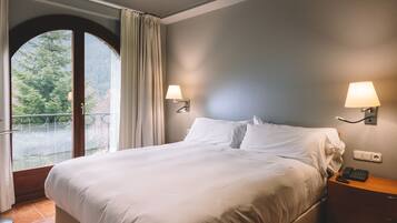Habitación de hotel con cama, dos lámparas fijadas a la pared, una mesita de noche, un teléfono y vistas a árboles a través de un ventanal grande.