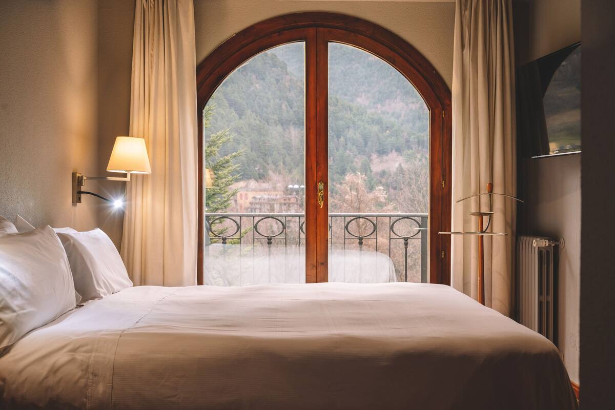A hotel room with a large bed, a view of a balcony, and a window with curtains.