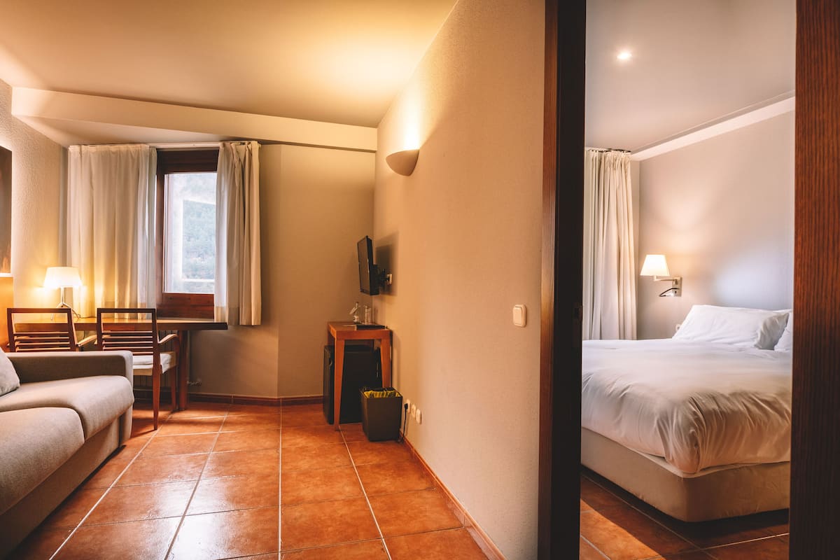 A hotel room with a bed, a sofa, a desk with a chair, a TV, and a trash bin.
