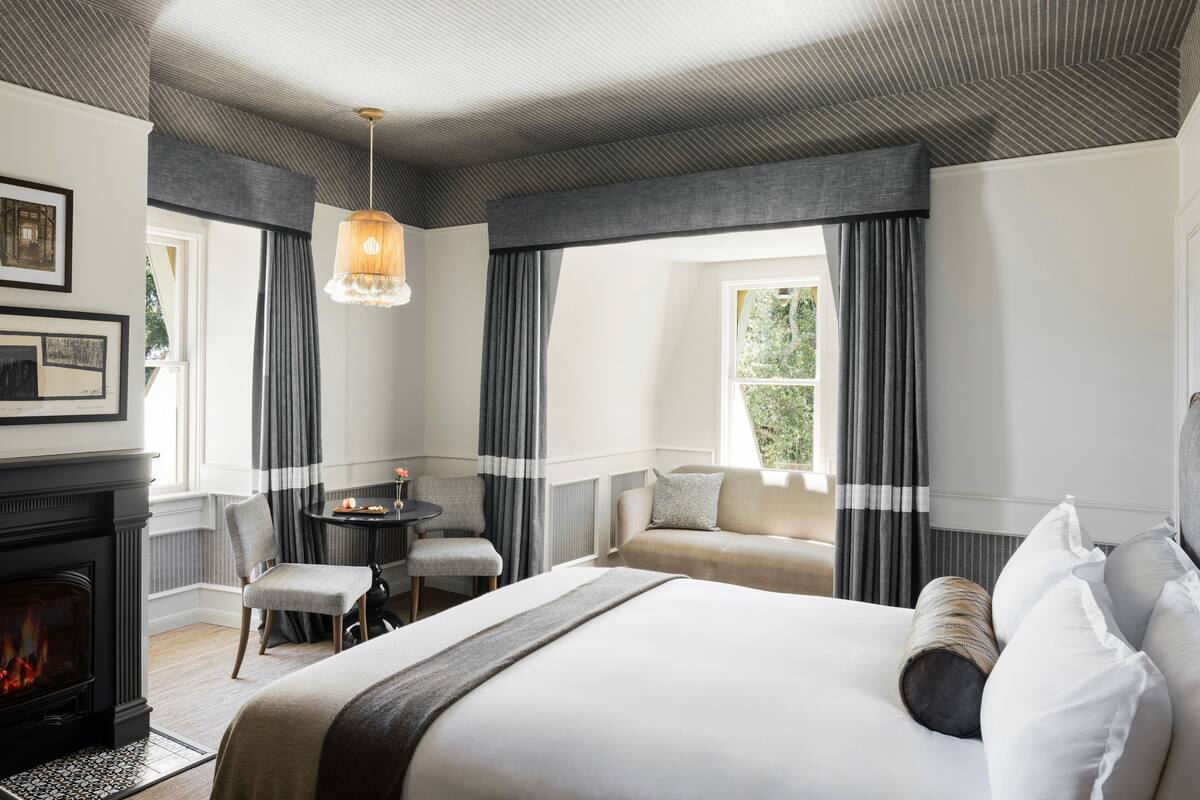 A hotel room with a large bed, a fireplace, a sofa, a dining table, and a view of the outdoors.