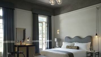 A spacious bedroom with a large bed, a desk, and a chair. There is a window with curtains, a chandelier, and a bedside table with a lamp.