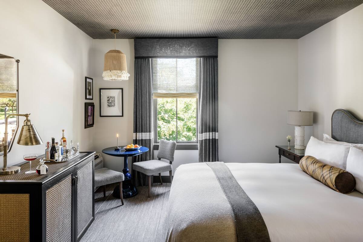 A hotel room with a large bed, a desk with a lamp, a chair, a small table, and a window with curtains.