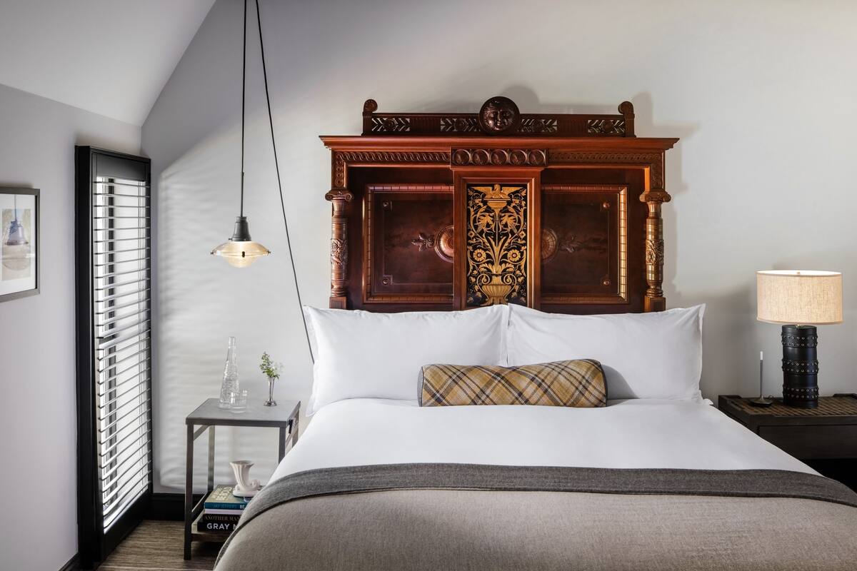 A hotel room with a large bed, a wooden headboard, a bedside table with a lamp, and a window with blinds.