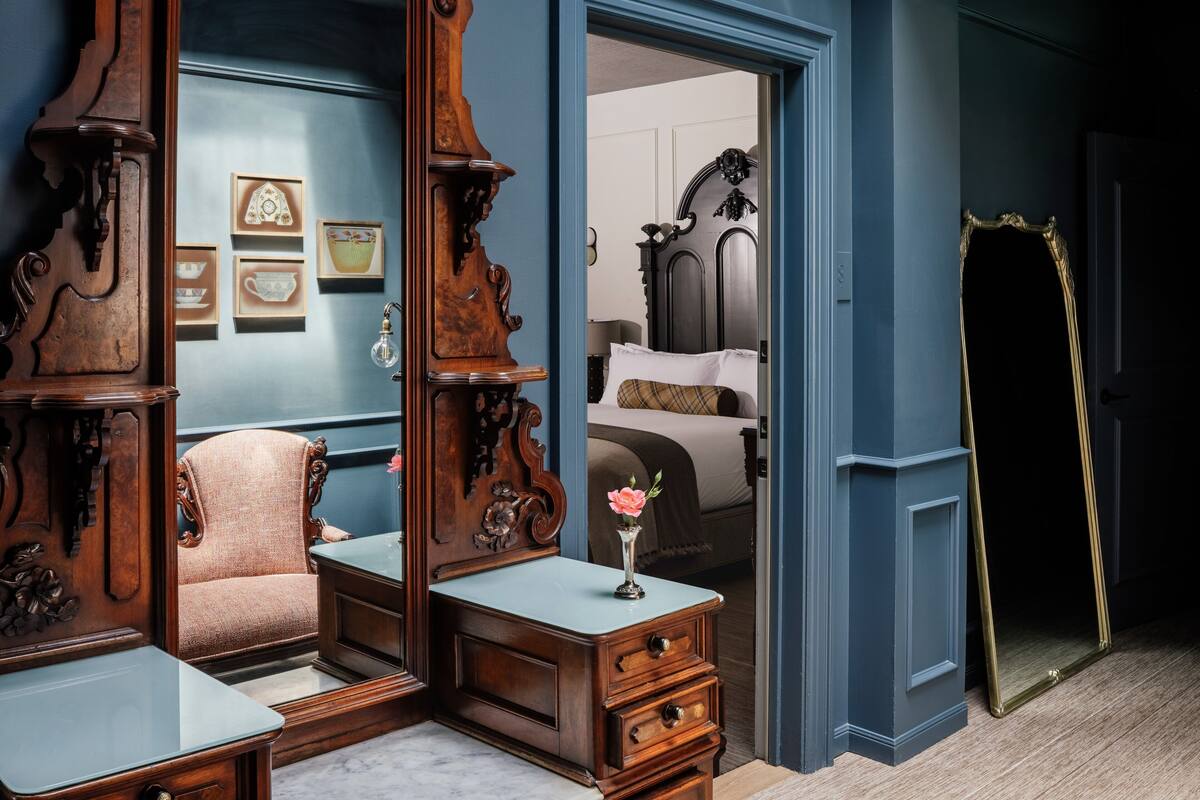 A bedroom with a large mirror, a wooden dresser, a chair, and a bedside table with a vase of flowers.
