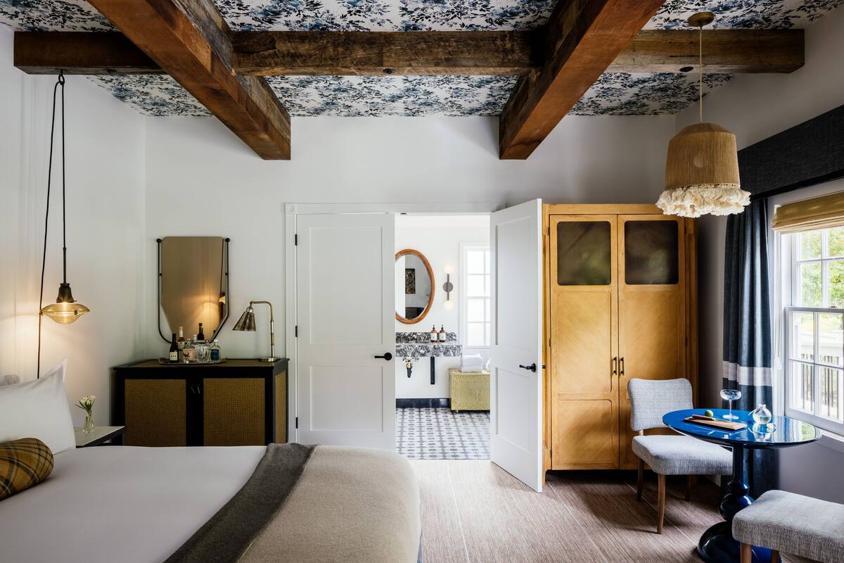 A hotel room with a bed, a desk with a lamp, a wardrobe, a dining table with chairs, and a view of the hallway.