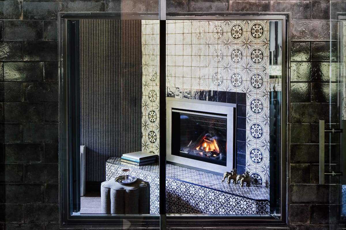 A bathroom with a large mirror reflecting a fireplace, a bathtub, and a tiled floor.