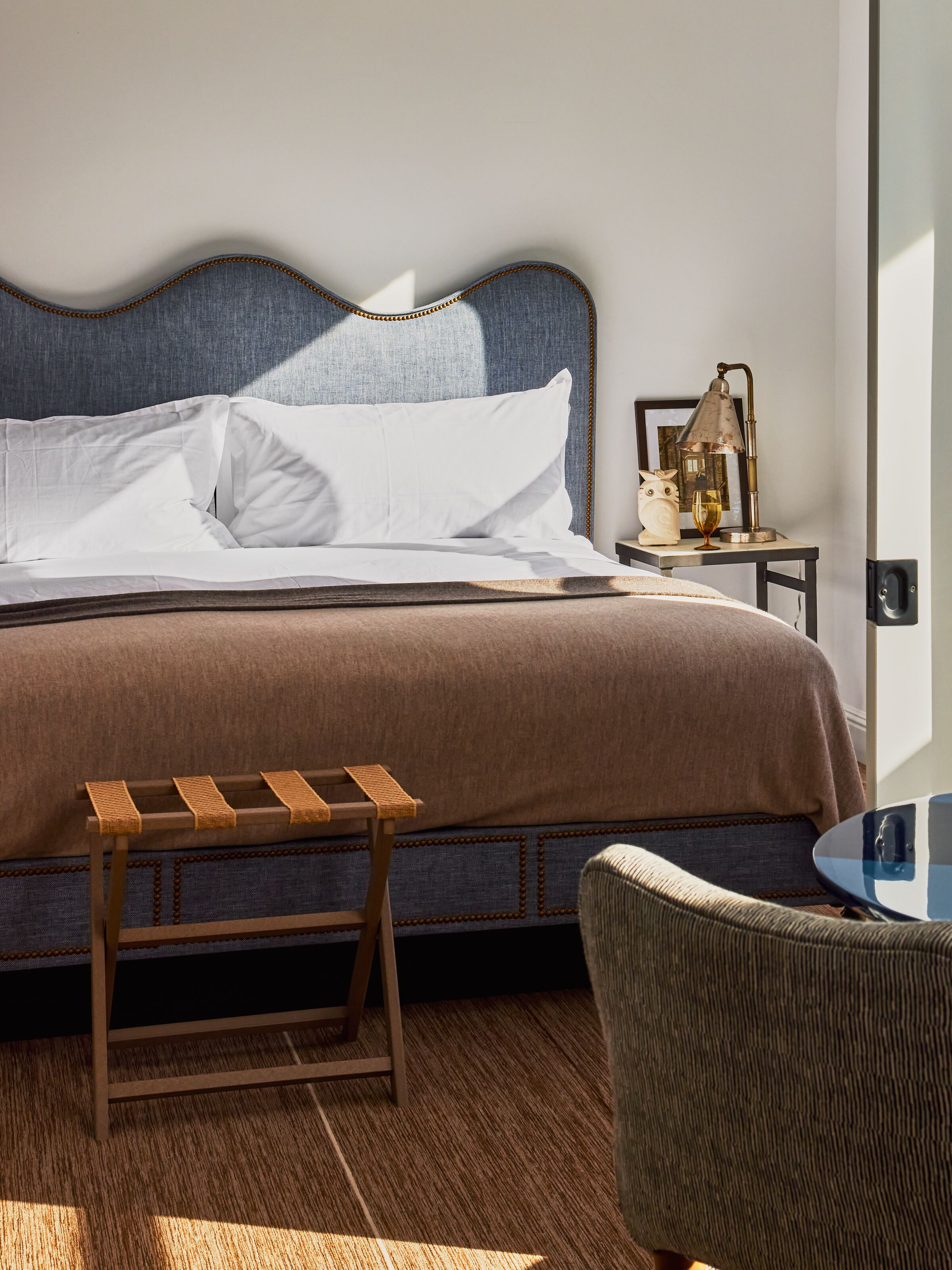 A neatly made bed with white linens, a wooden bench, and a small table with a lamp.