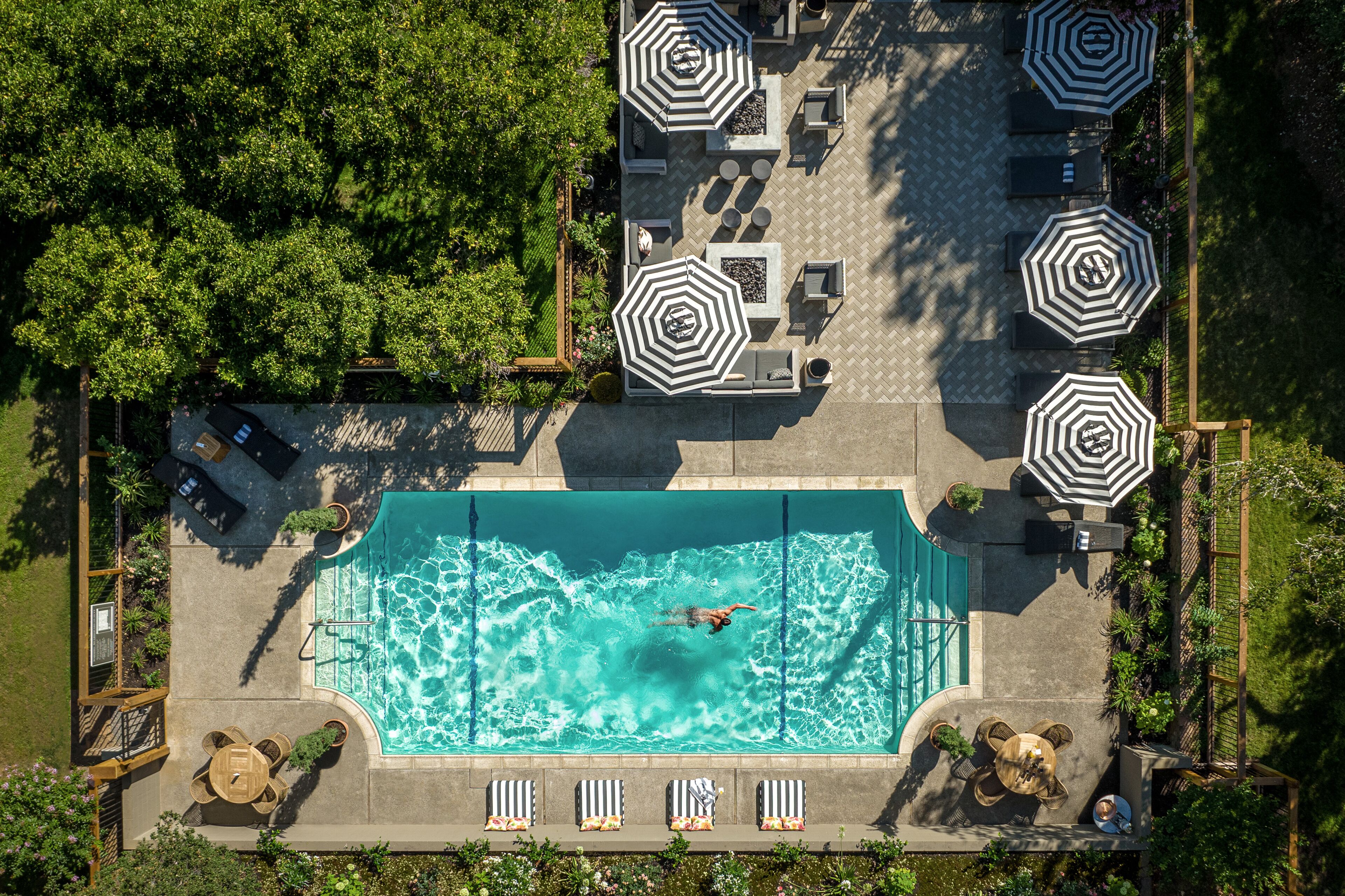 An aerial view of a swimming pool area with multiple lounge chairs and umbrellas, surrounded by lush greenery.
