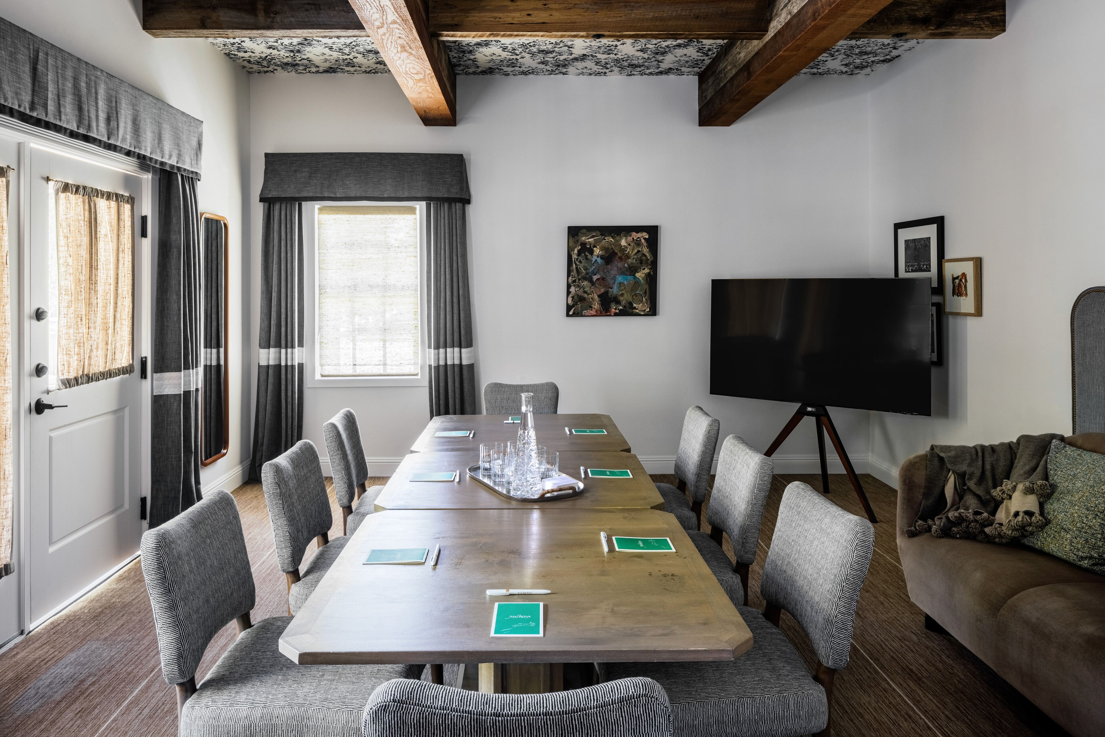 Ein Konferenzraum mit einem langen Tisch, grauen Polsterstühlen, einem großen Flachbildfernseher auf einem Stativ und einer Couch mit Decke und Kissen.