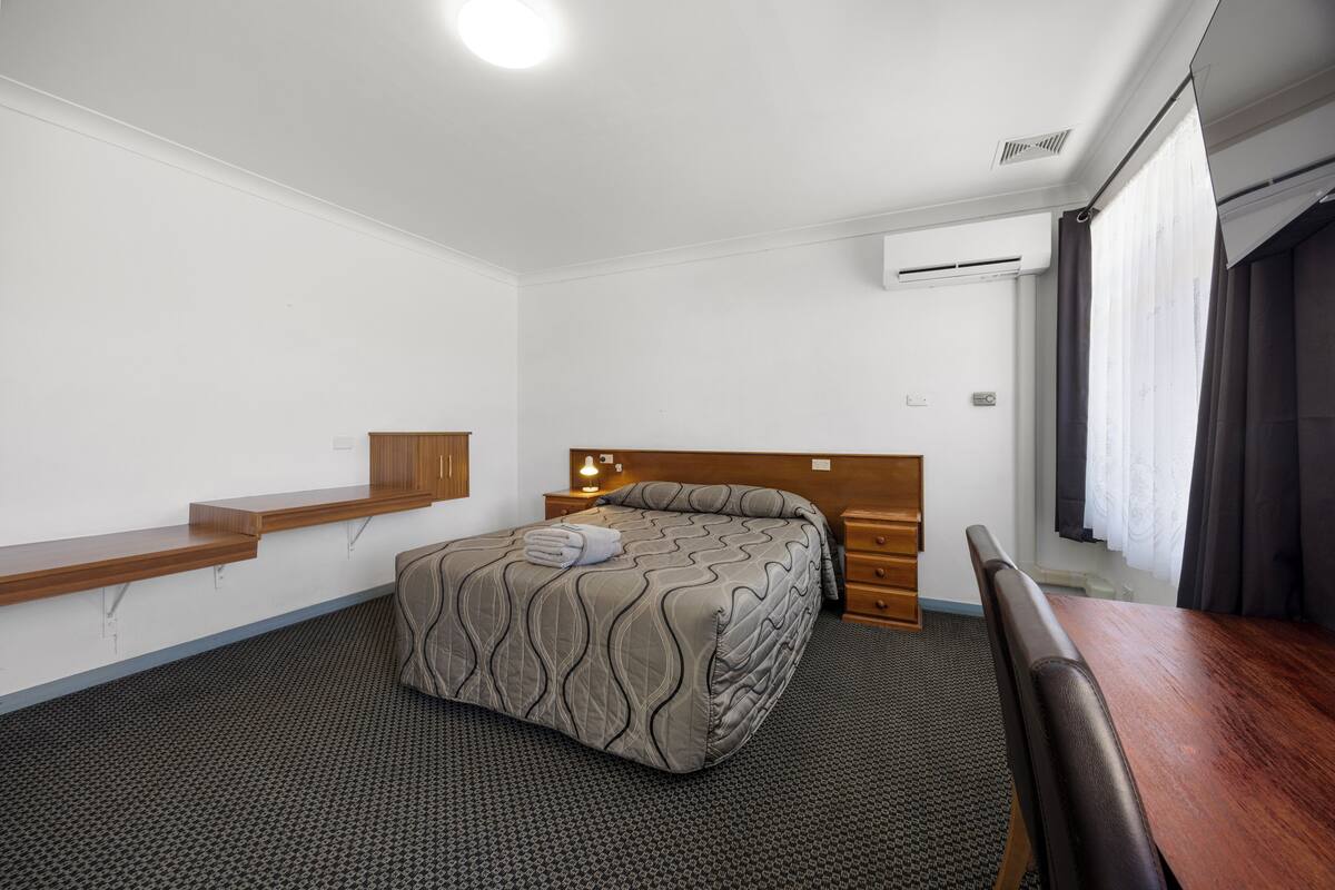 A hotel room with a bed, a wooden bench, a desk, and a window with curtains.