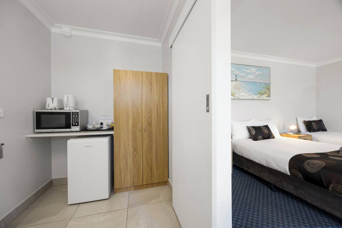 A compact hotel room with a kitchenette, a bed with patterned bedding, and a painting of a beach scene.
