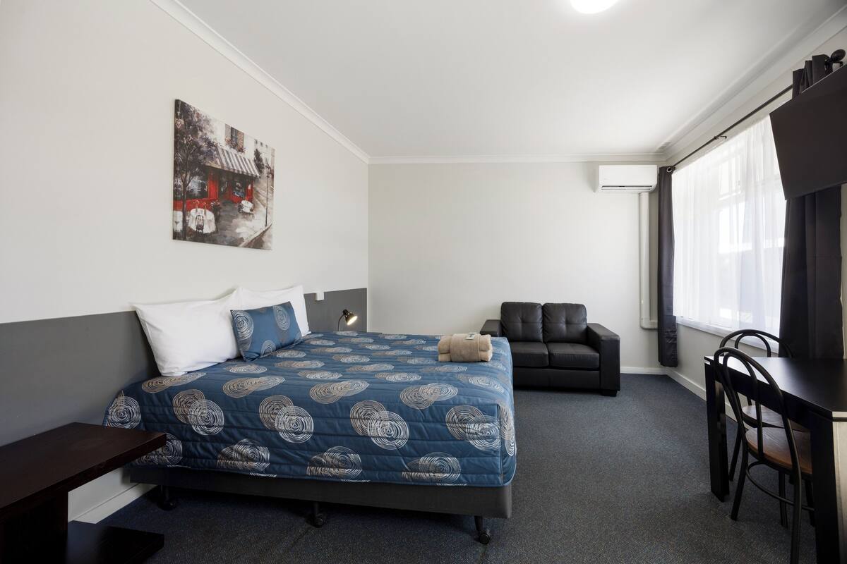 A bedroom with a bed, a desk, a chair, a sofa, and a painting on the wall.