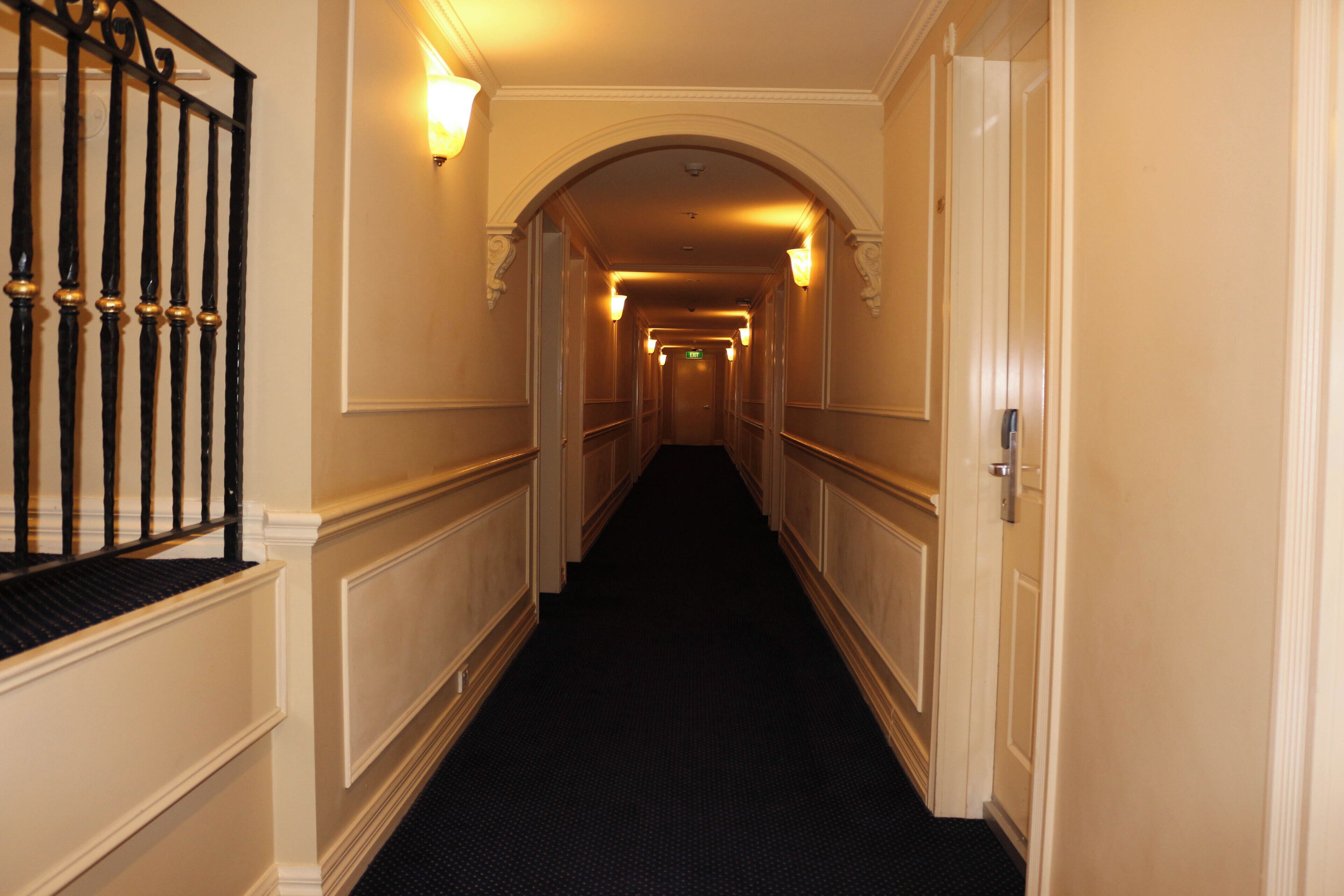 Un couloir d’hôtel avec une moquette sombre, des murs blancs et une rambarde en métal noir sur le côté gauche.