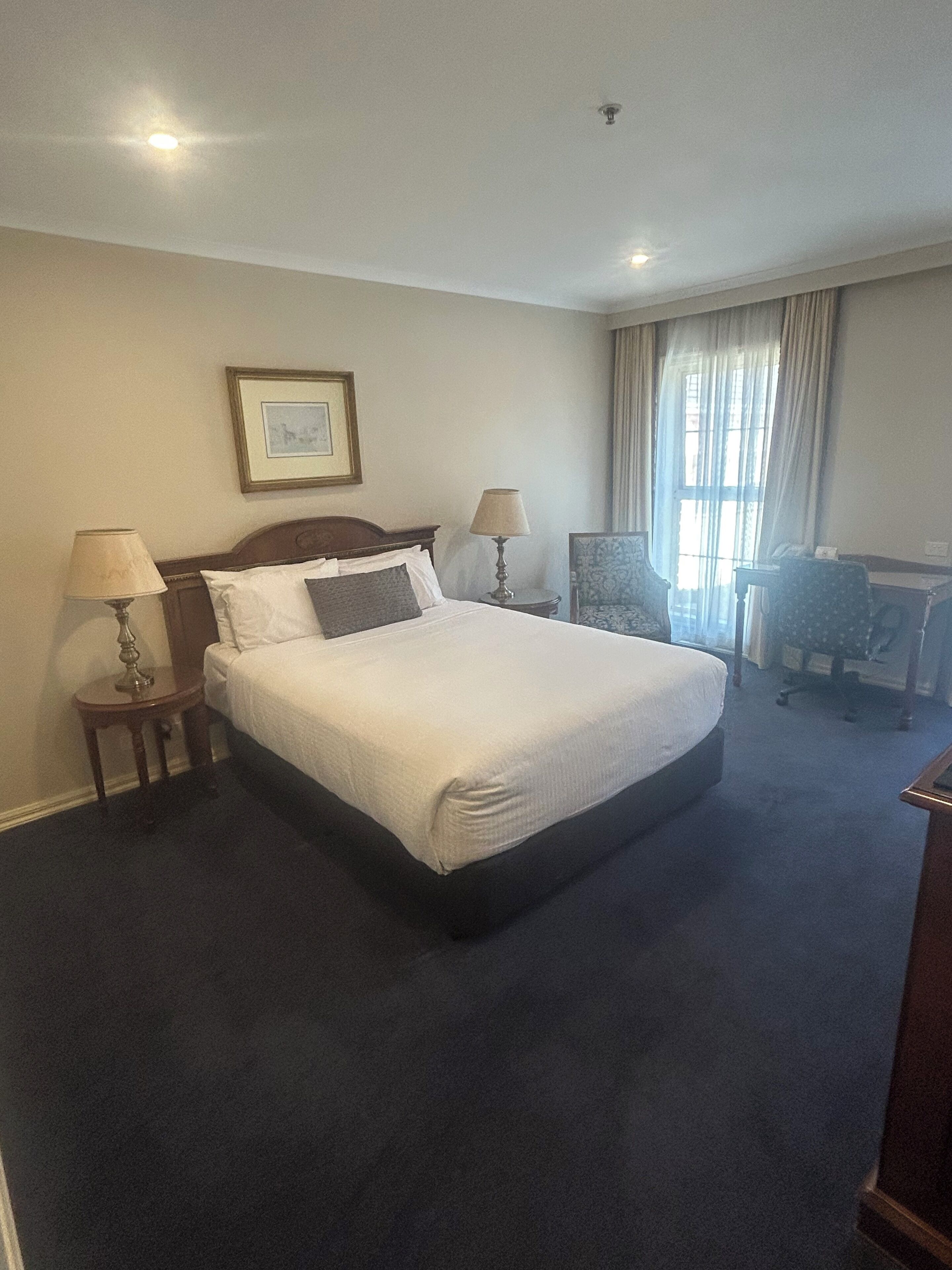 A hotel room with a large bed, two bedside tables with lamps, a chair, a desk, and a window with curtains.