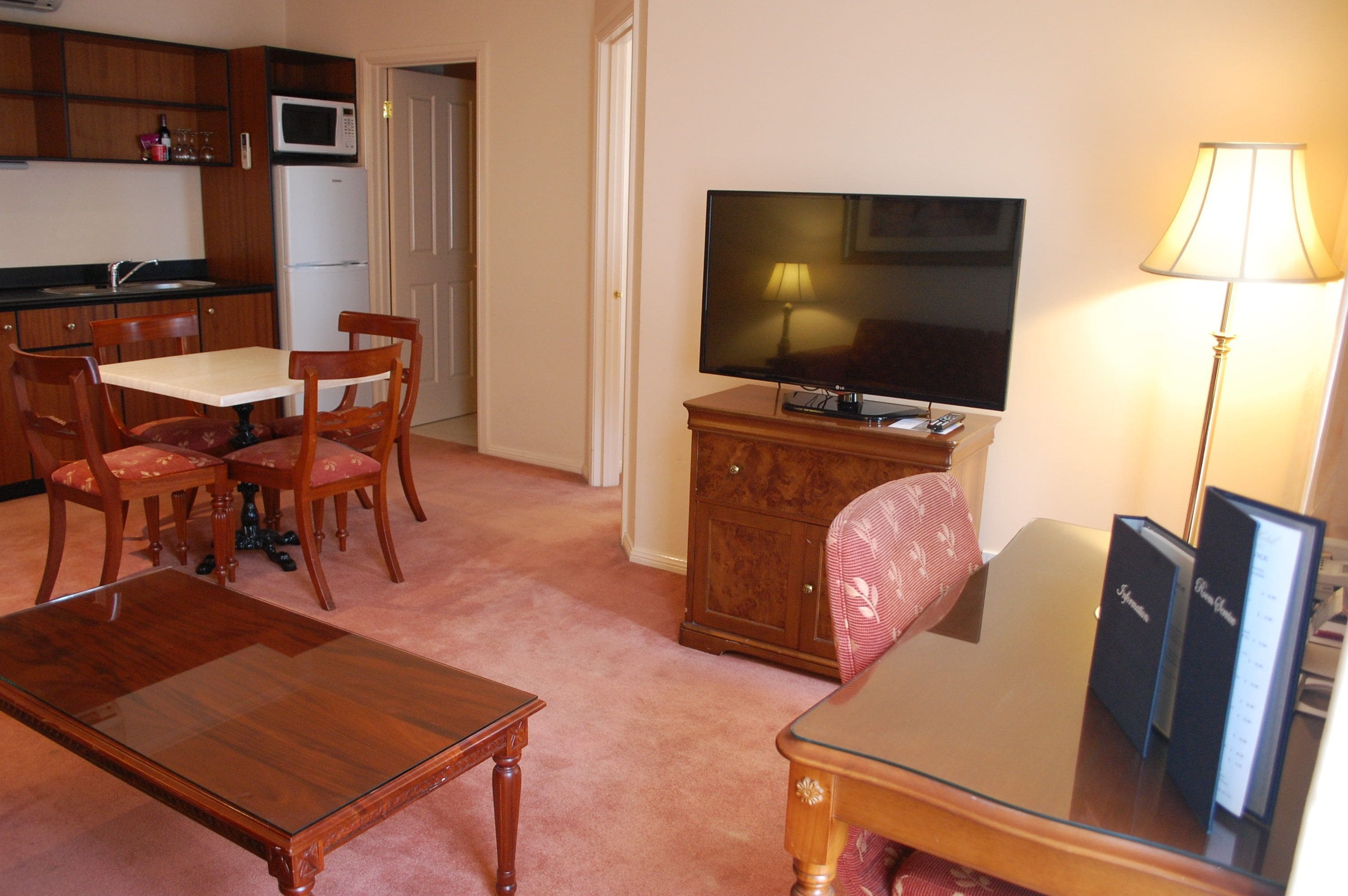 A living room with a wooden coffee table, a glass-top dining table with chairs, a flat-screen TV on a wooden cabinet, and a lamp.