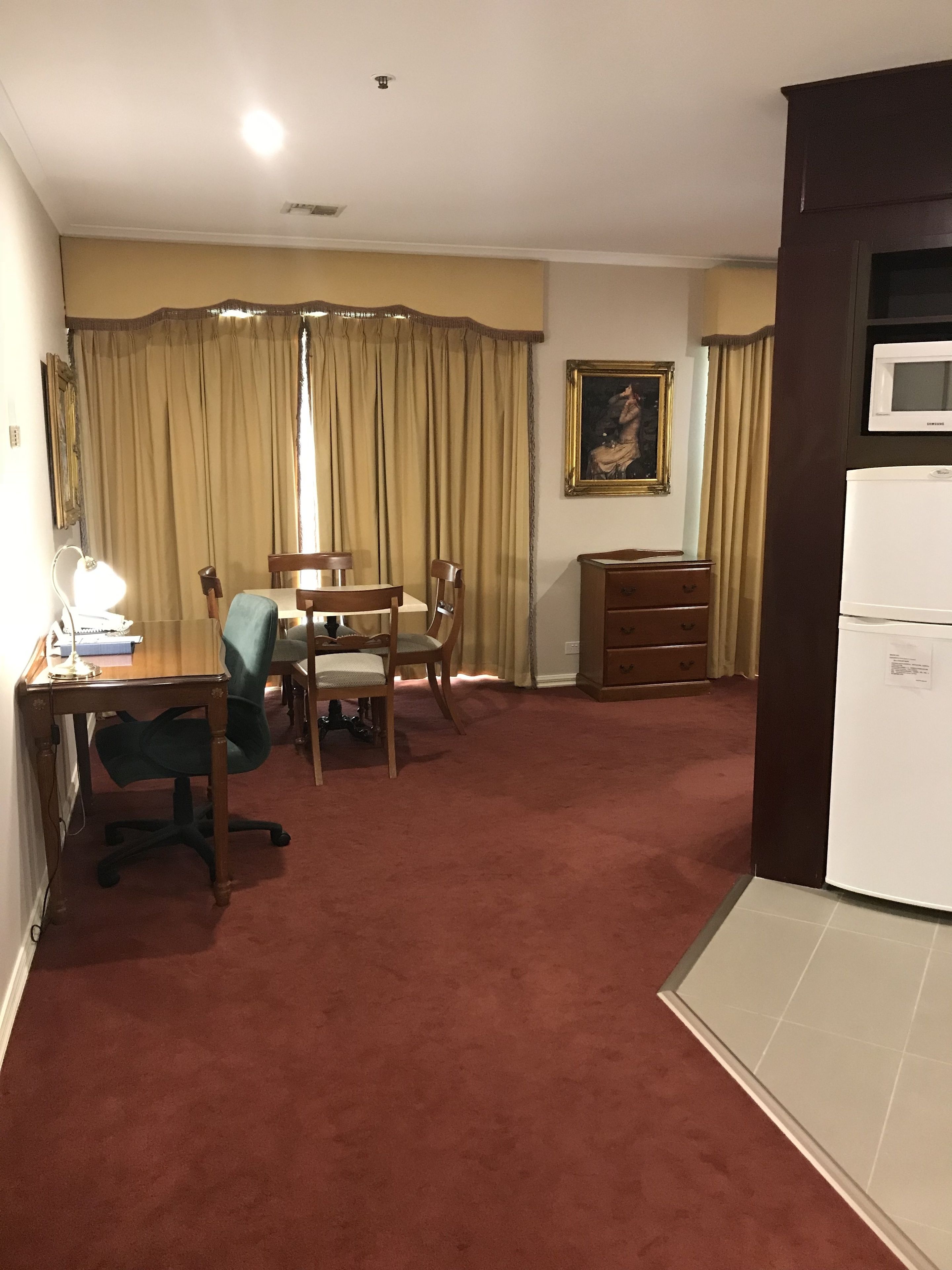 A hotel room with a dining area, a desk with a lamp, a chair, a small cabinet, and a microwave.