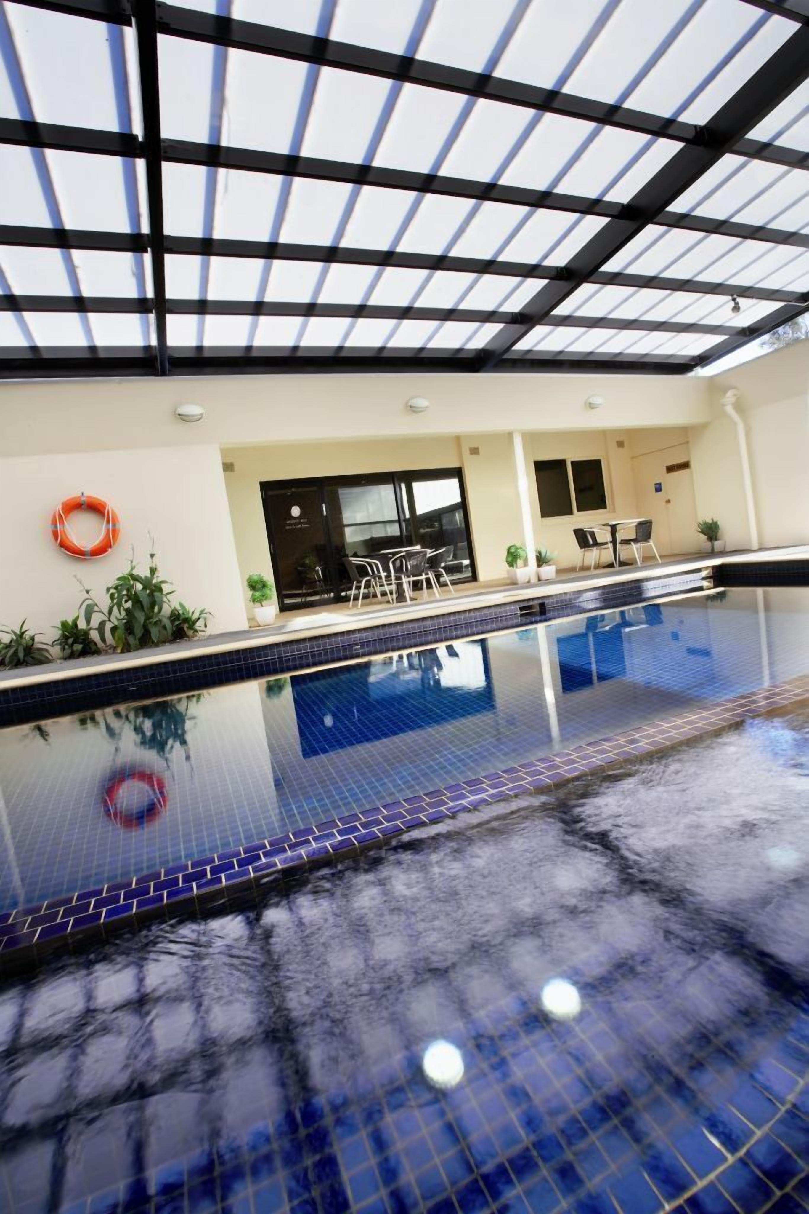 Una piscina interior con suelo de baldosas, techo de cristal y zona de estar con mesas y sillas.