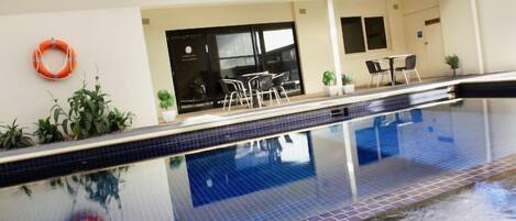 Una piscina interior con suelo de baldosas, techo de cristal y zona de estar con mesas y sillas.