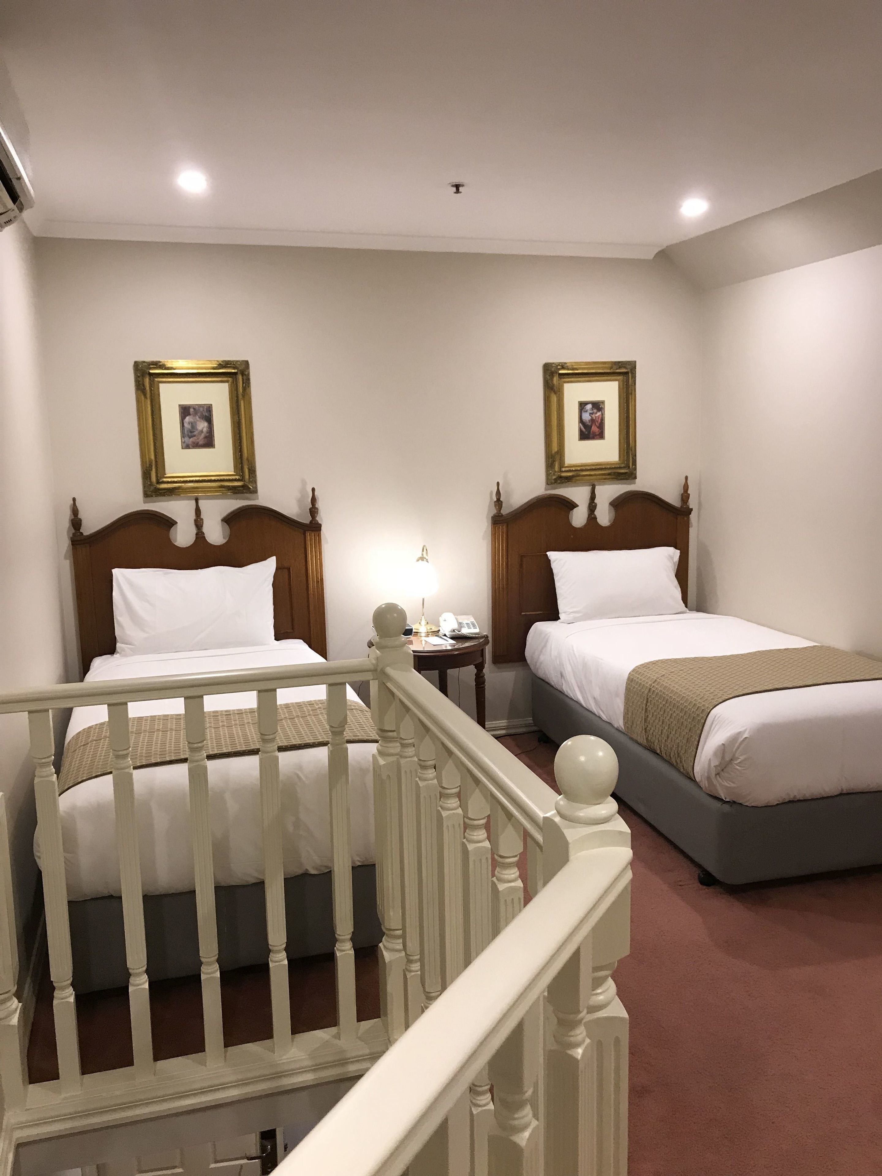 Habitación de hotel con dos camas individuales, una mesita de noche pequeña con lámpara y un cuadro en la pared.