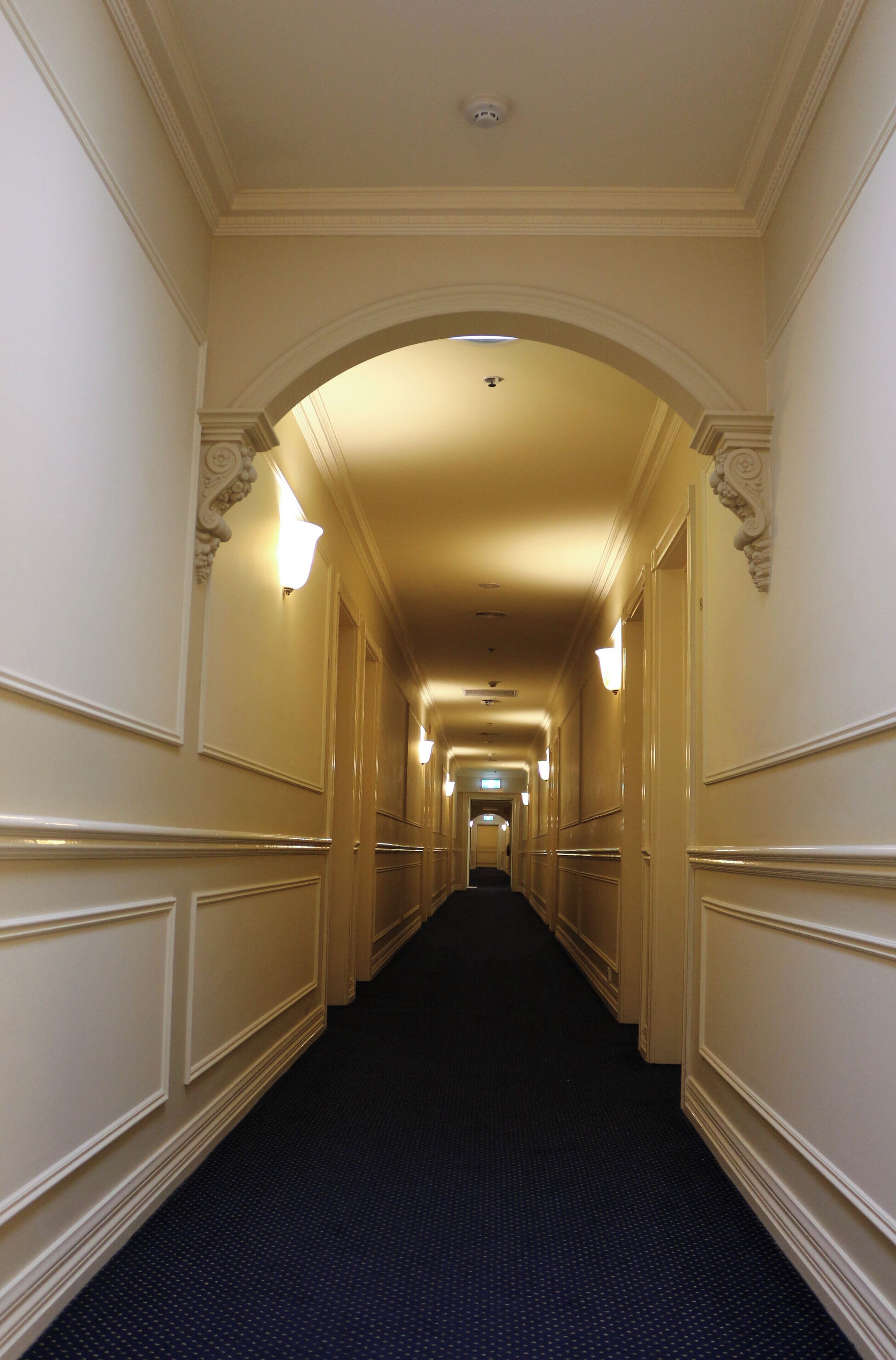 A long, narrow hallway with a dark carpet, white walls, and decorative moldings.