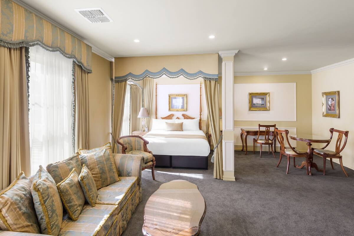 Habitación de hotel con una cama grande, un sofá, una mesa de comedor con sillas y un ventanal con cortinas.