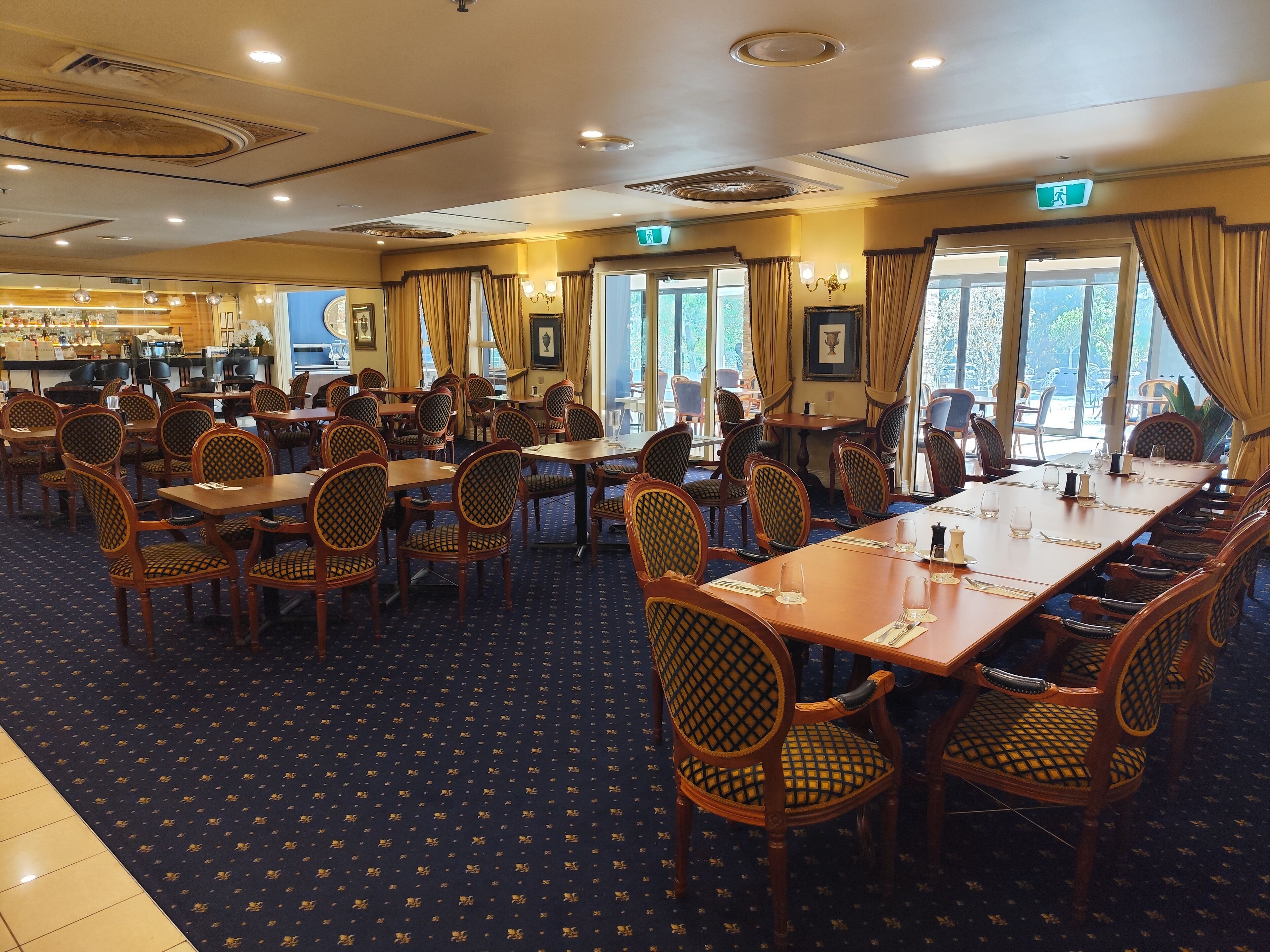 A hotel dining area with multiple tables and chairs, a bar in the background, and large windows with curtains.