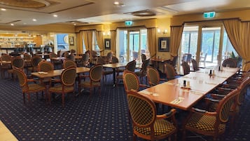 En hotelrestaurant med flere borde og stole, en bar i baggrunden og store vinduer med gardiner.