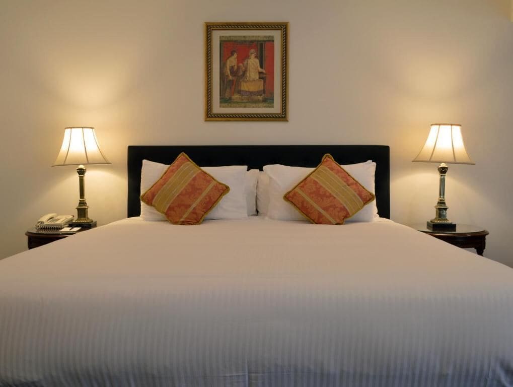 A neatly made bed with white linens, two decorative pillows, a framed artwork above, and bedside lamps on either side.