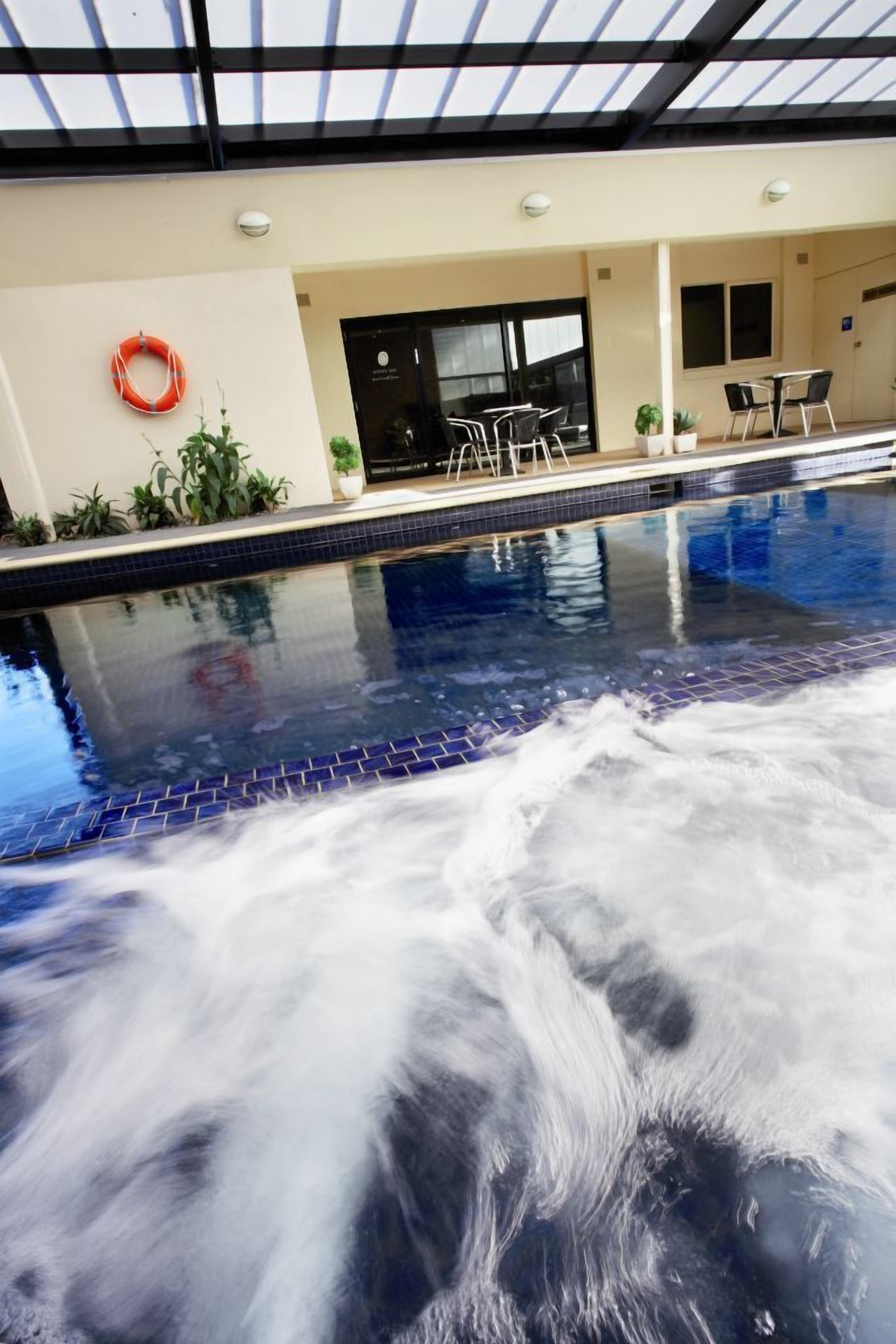 An indoor swimming pool with a tiled edge, a lifebuoy on the wall, and a seating area with chairs and tables.