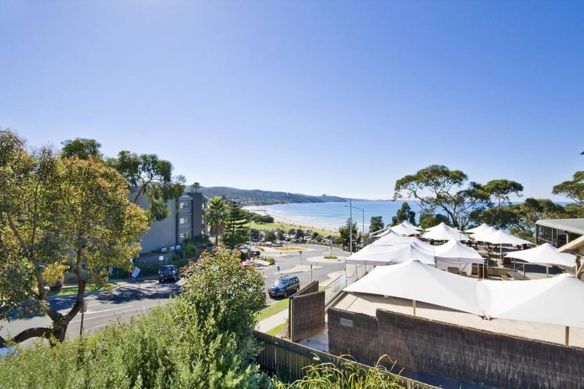 A coastal hotel with a view of the beach, a parking area, and outdoor seating under umbrellas.