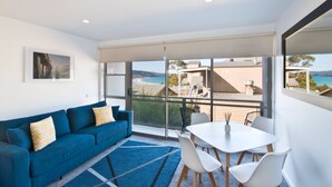 A modern living room with a blue sofa, white chairs, and a table. Large windows offer a view of the ocean and a building across the street. - Lorne Bay View Motel (Lorne)