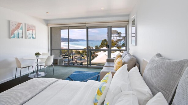 A modern hotel room with a large bed, a dining area with a table and chairs, and a balcony with a view of the beach and ocean. - Lorne Bay View Motel (Lorne)