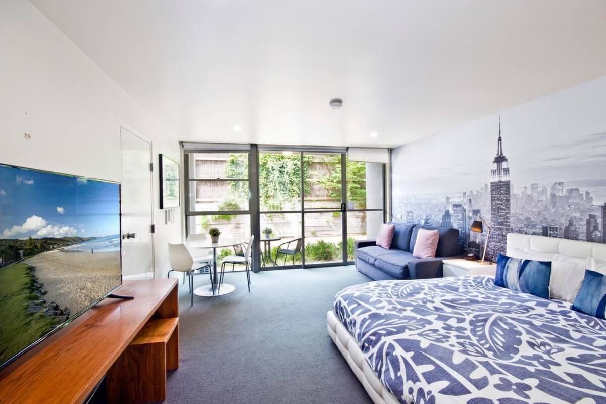 A modern bedroom with a large bed, a sofa, a desk, and a cityscape mural.