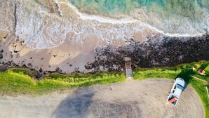 An aerial view of a beach with a rocky shoreline, a white van parked on the sand, and a small structure with a ladder leading to a rocky area. - Lorne Bay View Motel (Lorne)