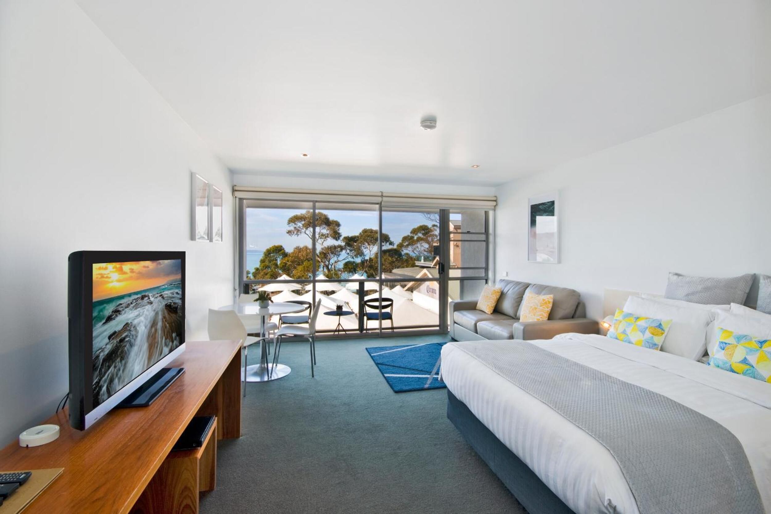 A modern hotel room with a large bed, a TV on a wooden desk, a balcony with outdoor seating, and a view of the city.