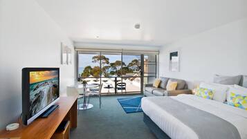 A modern hotel room with a large bed, a TV on a wooden desk, a balcony with outdoor seating, and a view of the city.