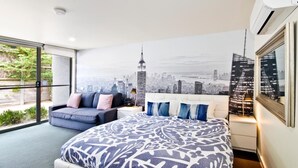 A modern bedroom with a cityscape mural, a large bed, a sofa, and a view of greenery outside. - Lorne Bay View Motel (Lorne)