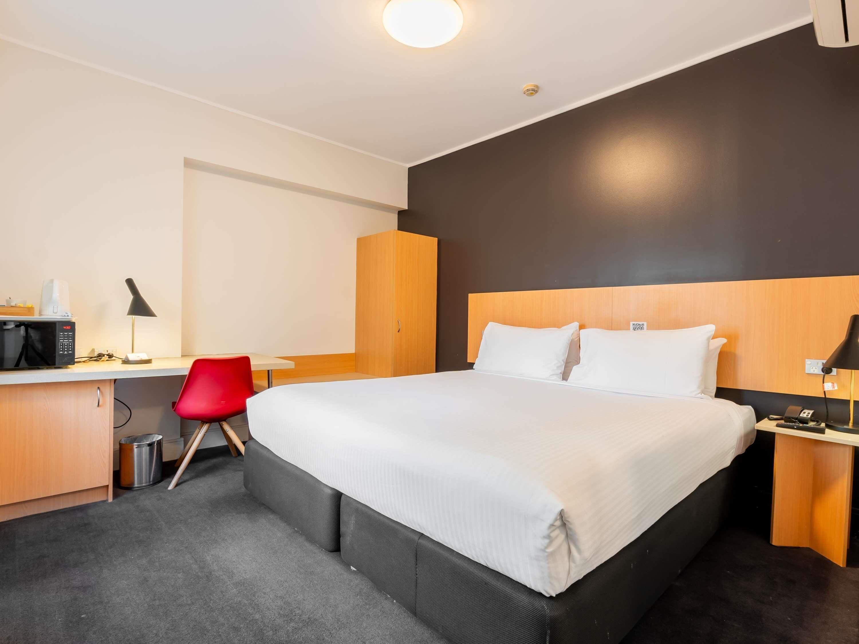 Ein Hotelzimmer mit einem Bett, einem Schreibtisch, einem roten Sessel, einem Kleiderschrank und einem Nachttisch mit Lampe.