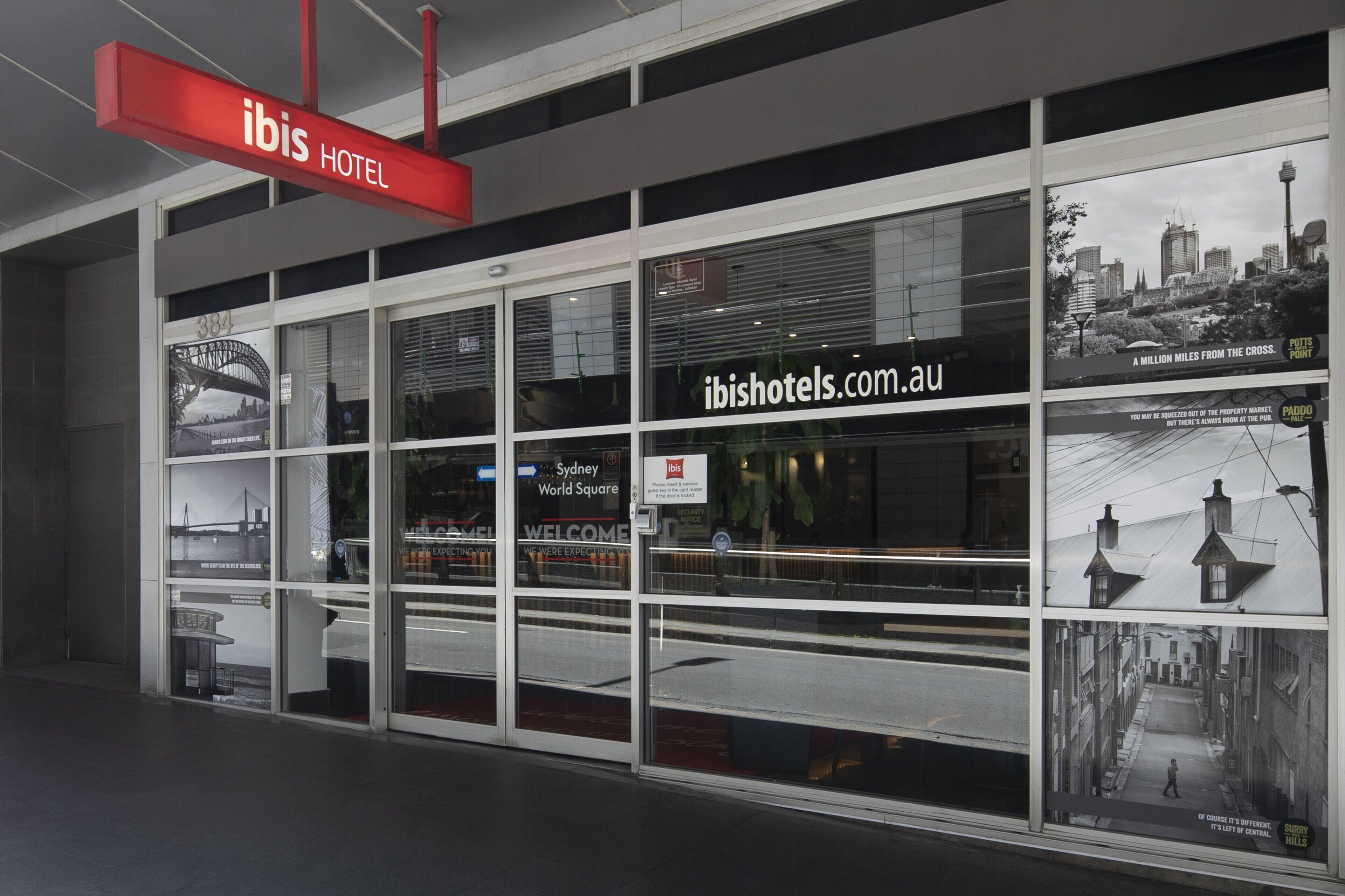 The exterior of the ibis hotel with a prominent red sign, large windows displaying the Sydney World Square view, and a website address on the window.