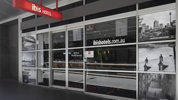 The exterior of the ibis hotel with a prominent red sign, large windows displaying the Sydney World Square view, and a website address on the window.